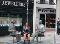 Eine junge Frau hat in Galway (Irland) zwischen zwei Bronze-Skulpturen Platz genommen.